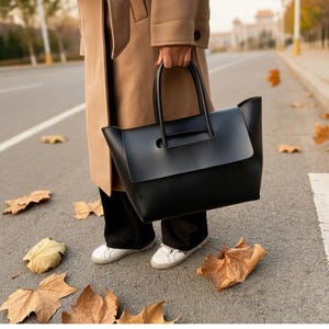 May include: A large black structured tote bag with a wide flap closure and two rounded top handles. The bag features a smooth matte finish and a rectangular silhouette, held by a hand at the side, showcasing a clean and functional design.