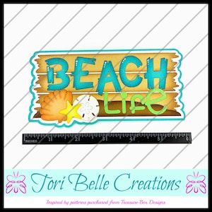 May include: A decorative sign with the words "BEACH Life" in turquoise and green, set against a wooden plank background. The sign includes seashell and starfish accents. A ruler at the bottom measures up to 6 inches.