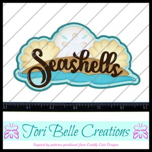 May include: A brown paper cut-out of the word "Seashells" with a blue wave and a white seashell on top. The design is inspired by patterns purchased from Cuddly Cute Designs.