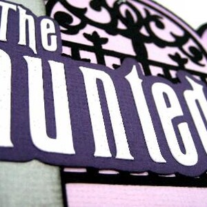 May include: The Haunted Mansion sign with white lettering on a purple background. The sign is framed by a black, ornate, gate-like design.