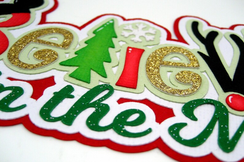 Puede incluir: Decoraci&oacute;n navide&ntilde;a festiva con la frase "Believe in the Magic of Christmas". Las letras son de purpurina dorada, verde, roja y blanca, con detalles de &aacute;rbol de Navidad y copo de nieve.