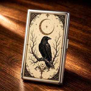 May include: A rectangular metal cigarette case featuring a cream-colored front panel with a black ink illustration of a raven perched on a bare branch under a crescent moon. The design includes ornate corner flourishes and a polished silver frame.