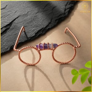 Könnte beinhalten: Dekorative Brille aus gedrehtem Kupferdraht mit einem horizontalen, violetten Amethyst-Kristallpunkt in der Mitte. Der Rahmen besteht aus zwei kreisförmigen Drahtringen, die durch den Kristall verbunden sind, mit zwei seitlichen Bügeln.