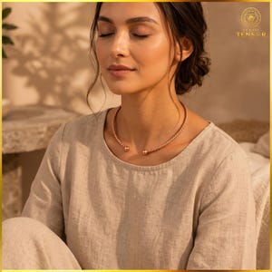 May include: A rose gold-toned metal torque necklace featuring a twisted rope-like texture and two small spherical end caps. The necklace is worn around the neck, resting against a light beige linen-textured long-sleeved top with a round neckline.