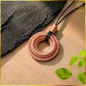 Può includere: Un ciondolo in filo di rame caratterizzato da un design a bobina circolare realizzato con fili metallici intrecciati. Il ciondolo è sospeso a un cordino nero e include un piccolo connettore cilindrico in rame sulla parte superiore. Il testo DA NANG TENSOR è visibile.