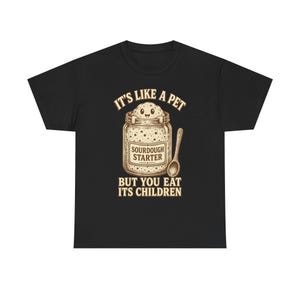 Könnte beinhalten: Ein schwarzes T-Shirt mit einer Grafik eines Glasgefäßes, das Sauerteigstarter enthält, aus dem ein lächelndes Wesen hervorschaut. Der Text lautet IT'S LIKE A PET BUT YOU EAT ITS CHILDREN und SOURDOUGH STARTER, ergänzt durch einen Holzlöffel.