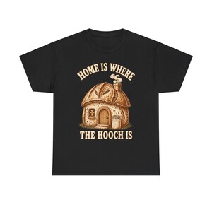 Könnte beinhalten: Ein schwarzes T-Shirt mit einer Grafik eines Hauses in Form eines Brotlaibs, inklusive rauchendem Schornstein, kleiner Tür und einem Glas daneben. Der beigefarbene Text über und unter der Illustration lautet "HOME IS WHERE THE HOOCH IS".