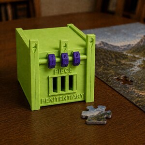 May include: A lime green plastic box shaped like a jail cell with three purple rotating number dials on the front. The word PIECE is embossed above a barred window, and the word PENITENTIARY is embossed below it. A single puzzle piece sits nearby.