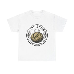 Könnte beinhalten: Ein weißes kurzärmeliges T-Shirt mit einer kreisförmigen Grafik auf der Brust. Das Design zeigt einen goldbraunen Sauerteiglaib mit Dampflinien, umgeben von Weizenähren und dem Text "LIFE IS HARD" sowie "GET A SOURDOUGH STARTER TO MAKE IT HARDER."