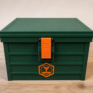 Könnte beinhalten: Eine rechteckige, dunkelgrüne Aufbewahrungsbox mit flachem Deckel und einem vertikalen, orangefarbenen Verschluss in der Mitte. Unter dem Verschluss befindet sich ein sechseckiges, orangefarbenes Emblem mit einem stilisierten Walschwanz-Symbol.