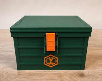 May include: A rectangular dark green storage box with a flat lid and a vertical orange latch centered on the front. Below the latch is a hexagonal orange emblem featuring a stylized whale tail icon. The box has horizontal recessed panel lines.