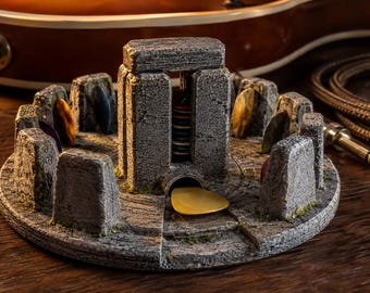 May include: A circular gray stone guitar pick holder designed like a miniature Stonehenge monument. It features upright stone pillars arranged in a circle around a central structure that holds a stack of colorful guitar picks, with one gold pick.