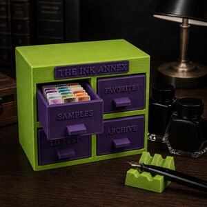Könnte beinhalten: Ein lindgrüner und violetter Aufbewahrungsschrank mit der Aufschrift THE INK ANNEX und vier Schubladen mit den Beschriftungen SAMPLES, FAVORITES, TO TEST und ARCHIVE. Die offene Schublade SAMPLES enthält kleine rechteckige Tintenproben. Inklusive zwei Tintenfässern.
