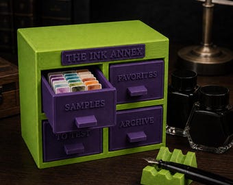 May include: A lime green and purple storage cabinet labeled THE INK ANNEX with four drawers labeled SAMPLES, FAVORITES, TO TEST, and ARCHIVE. The open SAMPLES drawer contains small rectangular ink swatches. Includes two black ink bottles and a pen rest.
