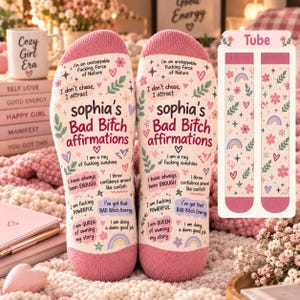 May include: A pair of light pink socks featuring the text 'sophia's Bad Bitch affirmations' alongside various phrases like 'I don't chase, I attract' and 'I am a ray of fucking sunshine', decorated with small hearts, flowers, and rainbows.