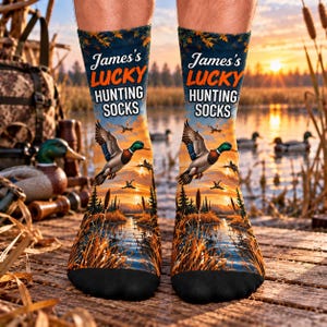 May include: A pair of crew socks featuring a printed sunset landscape with ducks flying over a marsh. The text reads James's LUCKY HUNTING SOCKS in white and orange lettering. The design includes tall grasses, trees, and water.