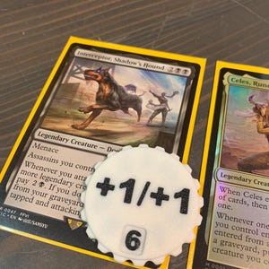 May include: A white, gear-shaped plastic counter token with black text reading +1/+1 and the number 6, placed on top of a Magic: The Gathering trading card titled Interceptor, Shadow's Hound, which features an illustration of a dark hound.