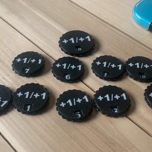 May include: A collection of ten black circular game counters with serrated edges, each featuring the white text +1/+1 and a small numerical display window at the bottom showing various digits ranging from zero to nine for tracking game statistics.