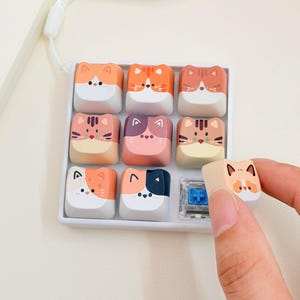 May include: A square white tray containing nine individual mechanical keyboard keycaps, each featuring a unique cat face design in various colors including orange, brown, pink, and black. One additional cat-themed keycap is held separately.