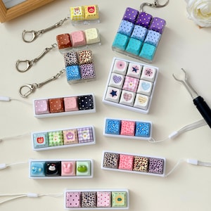 May include: A collection of mechanical keyboard keycap keychains and fidget toys featuring various designs including leopard prints, polka dots, hearts, stars, and animal faces in colors like blue, pink, purple, brown, and yellow with metal rings.