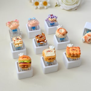 May include: A collection of mechanical keyboard keycaps featuring miniature food-themed characters, including a burger, toast, and cookies, each mounted on a square white base with colorful, detailed designs and small chef hats.