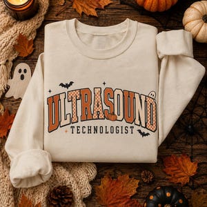 May include: A cream-colored crewneck sweatshirt featuring the text ULTRASOUND TECHNOLOGIST in orange and white checkered block letters. The design includes small black bat silhouettes, a tiny orange pumpkin, and four-pointed star accents.