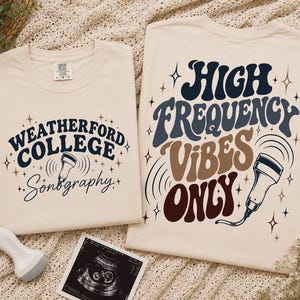 May include: Two cream-colored t-shirts featuring dark blue and brown graphic prints. The smaller shirt displays "WEATHERFORD COLLEGE Sonography" with a transducer icon. The larger shirt features "HIGH FREQUENCY VIBES ONLY" with a transducer.