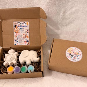 May include: Two brown cardboard boxes containing a craft kit. One open box reveals a white ceramic turtle and elephant, four small paint pots in orange, purple, green, and blue, and a Lunita Workshop instruction sticker on the inside lid.