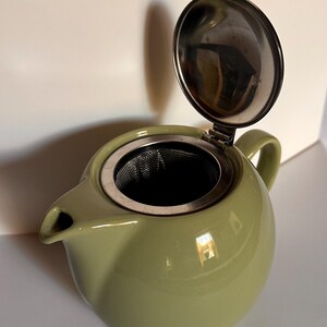 May include: A sage green ceramic teapot with a rounded body, a short spout, and a handle. The teapot features a hinged stainless steel lid that is open, revealing a removable fine-mesh metal tea infuser basket seated inside the opening.
