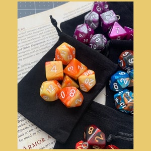 May include: A set of orange, yellow, and white dice with black numbers in a black velvet bag. There are also purple, blue, and red dice with white numbers.