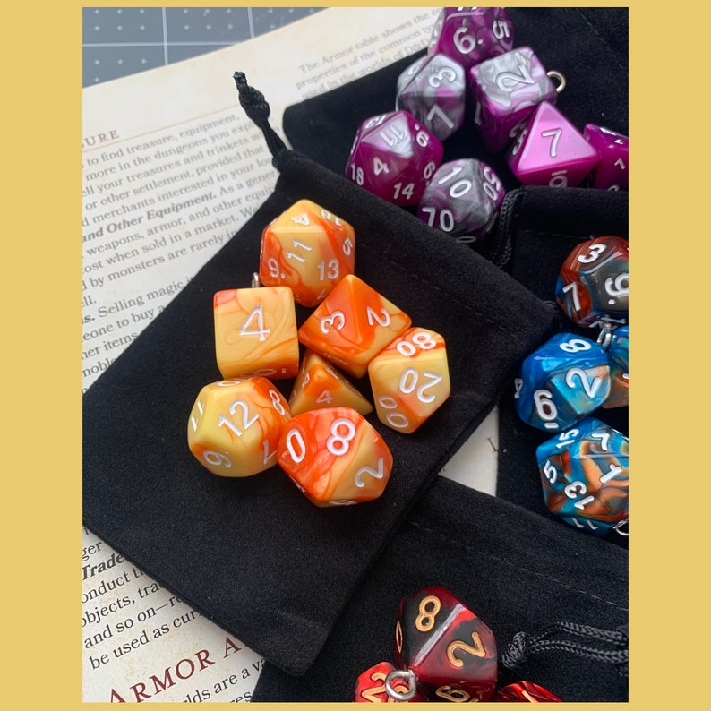 May include: A set of orange, yellow, and white dice with black numbers in a black velvet bag. There are also purple, blue, and red dice with white numbers.