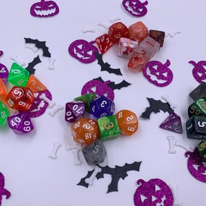 Peut inclure: Un assortiment de dés colorés, orange, vert, rouge et violet, dispersés sur une surface blanche. Les dés sont entourés de décorations sur le thème d'Halloween, comme des citrouilles violettes à paillettes, des chauves-souris noires et des confettis en forme d'os.