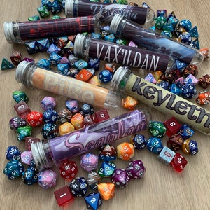 May include: A collection of six colorful, labeled test tubes filled with various multi-colored dice. The test tubes are labeled with the names Vex'ahlia, Vax'ildan, Pike, Keyleth, Scanlan, and Percival de Rolo III.