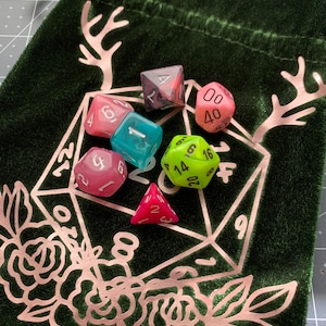 May include: A green velvet dice bag with a pink floral design and a pair of antlers. The bag contains six dice in various colours, including pink, green, blue, and black.