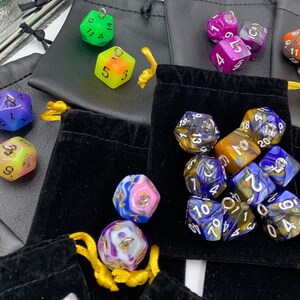 May include: A collection of colorful dice in various shapes and sizes, including a set of blue and gold dice with white numbers, a set of yellow and blue dice with white numbers, and a set of green and white dice with black numbers. The dice are arranged in a variety of patterns and are displayed on a black velvet surface.