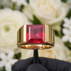 May include: A gold ring featuring a large, square-cut, vibrant red gemstone set in a polished yellow gold band. The setting is smooth and wide, highlighting the deep crimson colour and faceted surface of the central stone against a metallic finish.