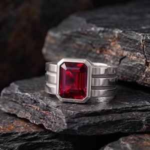 Puede incluir: Un anillo de tono plateado con una gran piedra preciosa de color rojo intenso, corte esmeralda rectangular y engaste de bisel. La banda está formada por tres capas horizontales paralelas, lisas y distintas, creando un perfil ancho.