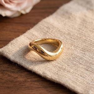 May include: A polished gold ring with a smooth, continuous wave-like design. The band features a thick, rounded profile with a high-shine reflective surface that curves gently, creating an organic, fluid shape that encircles the finger.