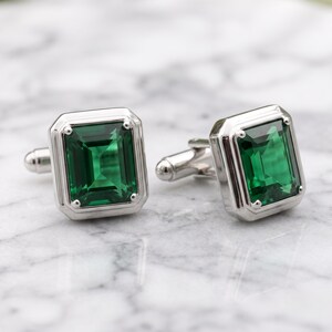 May include: A pair of silver-toned cufflinks featuring rectangular emerald green stones set in a polished metal frame. Each cufflink has a faceted, deep green center stone with a stepped border design and a standard hinged toggle closure mechanism.