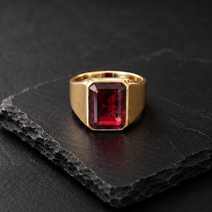Könnte beinhalten: Ein Goldring mit einem großen, rechteckigen, facettierten tiefroten Edelstein, der in eine glatte, polierte Goldschiene gefasst ist. Der Ring hat eine massive, solide Konstruktion mit einer breiten Ringschiene, die sich leicht verjüngt.