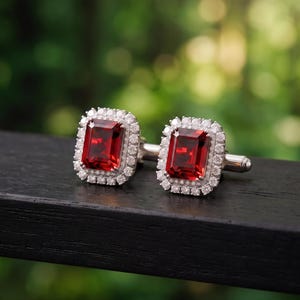 Puede incluir: Un par de gemelos de tono plateado, cada uno con una piedra preciosa roja profunda de corte rectangular esmeralda en el centro, rodeada por un halo de pequeñas piedras blancas de corte redondo engastadas en un marco metálico con cierre.