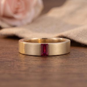 May include: A gold band ring featuring a single rectangular baguette-cut ruby gemstone set flush into the centre of the metal. The gold band has a brushed matte finish, providing a textured appearance that contrasts with the deep red gemstone.