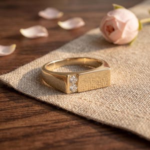 May include: A gold ring featuring a flat, rectangular top surface and a vertical channel set with two small, clear, round-cut stones. The band has a polished, smooth finish and a solid, wide construction that tapers toward the back of the ring.