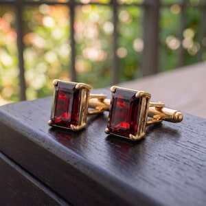 May include: A pair of gold cufflinks featuring rectangular, emerald-cut deep red garnet gemstones. Each stone is securely held by four gold prongs on a polished gold metal base with a standard toggle closure mechanism for fastening.