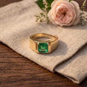 Peut inclure: Une bague chevalière en or ornée d'une pierre précieuse verte vibrante de taille carrée au centre. L'anneau est lisse et poli, présentant une construction large en métal massif qui encadre solidement la pierre verte à facettes.