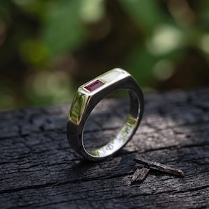 May include: A polished silver-toned metal ring featuring a single rectangular, deep red gemstone set horizontally into the flat top surface. The band has a smooth, solid construction with a reflective finish and a clean, geometric design profile.