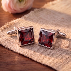 May include: A pair of square cufflinks featuring deep red, faceted stones set in polished silver-toned metal frames. Each cufflink has a hinged toggle mechanism on the back, designed to secure through shirt cuffs with a classic, clean aesthetic.