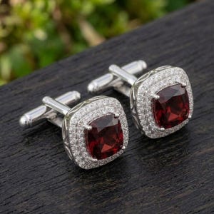 Puede incluir: Un par de gemelos de tono plateado con una piedra preciosa granate de color rojo intenso y corte cuadrado en el centro. Cada piedra está rodeada por un doble halo de pequeñas piedras claras y brillantes engastadas en pavé sobre una base metálica.