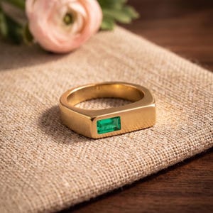 Peut inclure: Une bague dorée présentant un design de bande plate et rectangulaire avec une seule petite pierre précieuse verte de taille rectangulaire incrustée au centre de la surface supérieure. Le métal a une finition lisse et polie qui reflète la lumière.