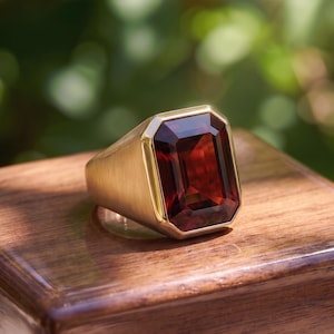 Può includere: Un anello in oro con una grande pietra preziosa rosso scuro di forma rettangolare a taglio smeraldo, incastonata in una lunetta in oro lucido. Il gambo dell'anello presenta una finitura spazzolata opaca che contrasta con la superficie sfaccettata.
